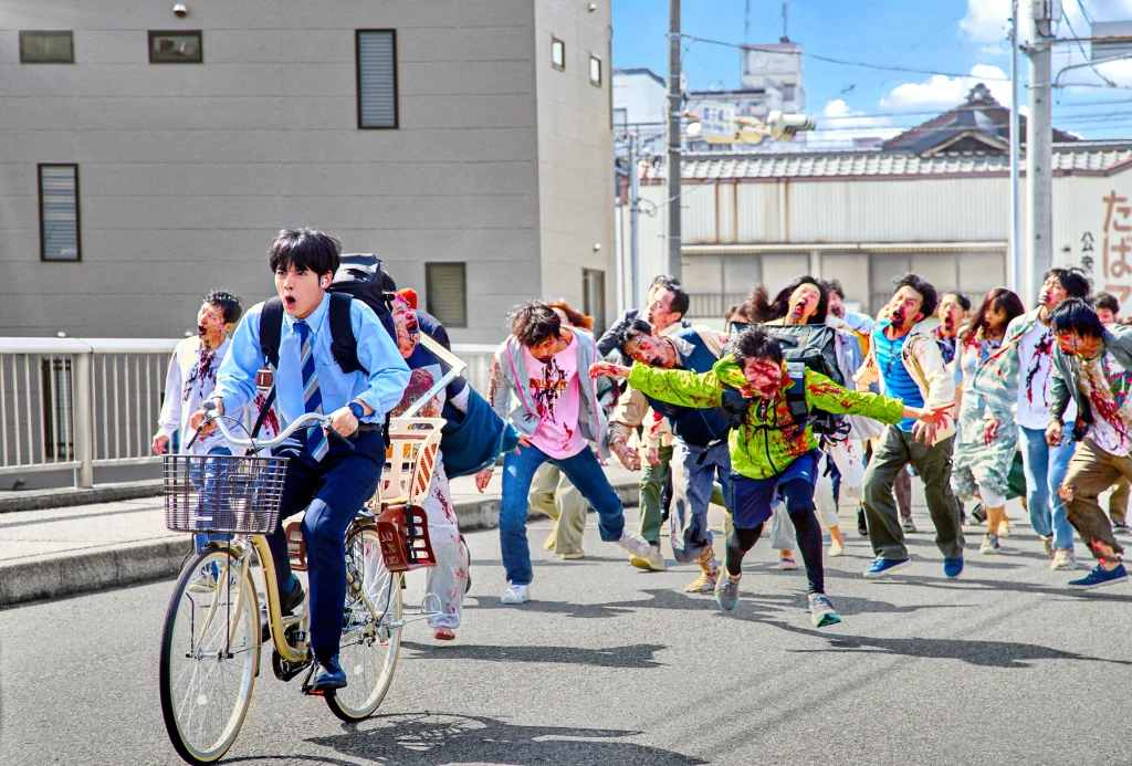 Akira (Eiji Akaso) hurridly cycles away from a swarm of pursuing zombies