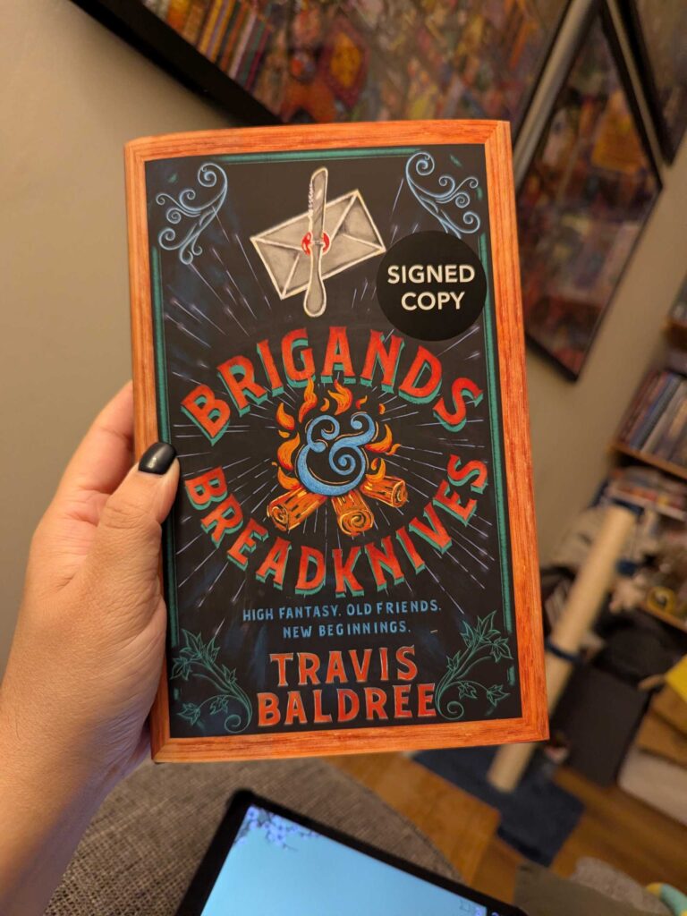 A photograph of the cover of Brigands & Breadknives by Travis Baldree. It has a sticker indicating its a signed copy. The tagline is "High fantasy, old friends, new beginnings."