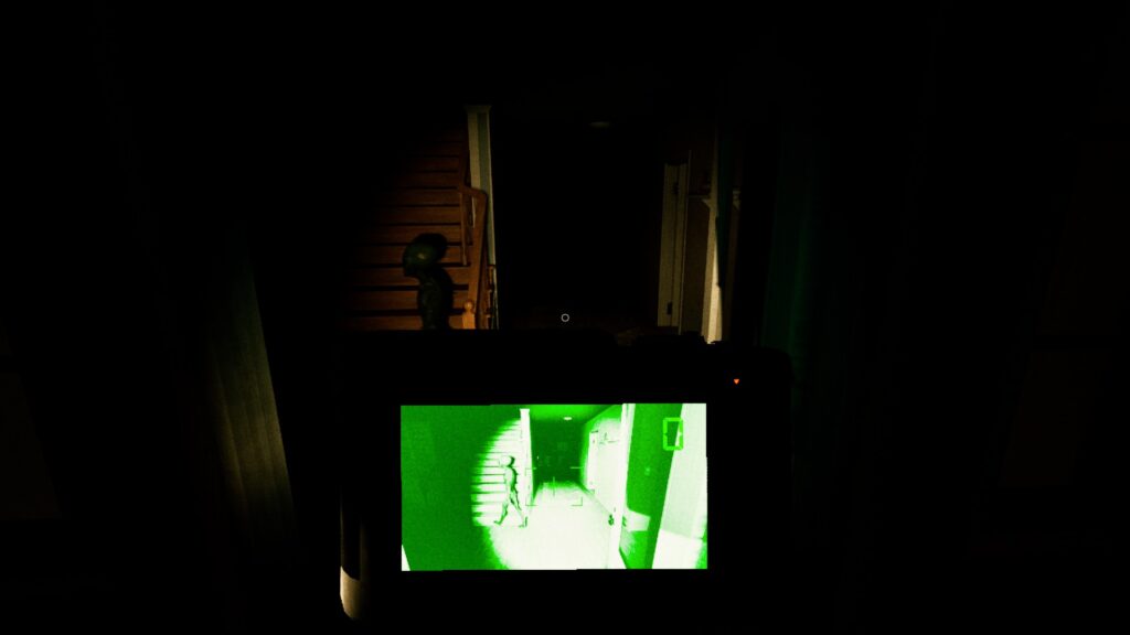 UFOPHILIA- A hallway to the right, and a wooden staircase to the left in a house. The only light is coming from a torch and the night vision of a camera at the bottom centre of the image. A dark green alien can be seen to the left of the screen facing the left as it goes towards the stairs. The camera is also showing the alien walking on its screen.
