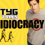 Yellow background with sun rays, a man to the right with a quizzical expression, arms raised outwards to his side, wearing a grey t-shirt with a red circle, the evolution of man in the bottom left corner, the name Idiocracy in the centre and TYG Talks logo just above the title to its right.